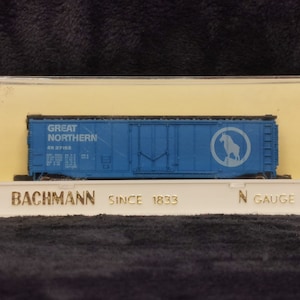 Może przedstawiać: Niebieski model wagonu towarowego z nadrukiem "Great Northern" i "GN 37163" na boku. Wagon ma również białe koło z kozłem górskim w środku. Wagon znajduje się w przezroczystej plastikowej gablocie z nadrukiem "Bachmann Since 1833 N Gauge" na dole.