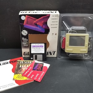 May include: A beige computer pointing device with a small screen and buttons. The device is packaged in a clear plastic case. The box for the device is white with blue and purple accents. The box has the text "Cirque Glidepoint Touchpad 2" on it. The device is accompanied by a floppy disk, a user manual, and a pamphlet.