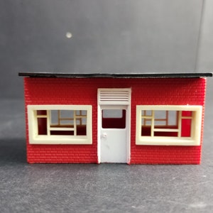 Puede incluir: Un edificio de ladrillo rojo con un techo negro, dos ventanas y una puerta blanca. El edificio es un modelo y está hecho de plástico.