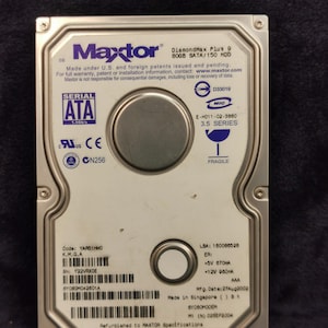 Puede incluir: Un disco duro plateado con el logotipo de Maxtor y el texto "DiamondMax Plus 9 80GB SATA/150 HDD". El disco duro tiene un conector SATA y una pegatina de frágil.