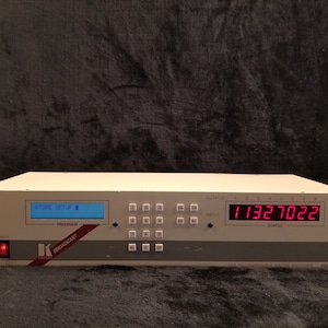 Puede incluir: Un mezclador de audio blanco con un botón de encendido rojo, una pantalla azul que muestra "STORE SETUP" y una pantalla digital roja que muestra "11327022". El mezclador tiene una fila de botones etiquetados como "PROGRAM" y "BROADCAST". El texto "K BROADCAST" es visible en el panel frontal. El texto "2000" es visible en la esquina inferior derecha del mezclador.