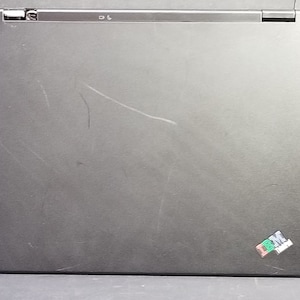 Może przedstawiać: Czarny laptop z srebrnym zawiasem i małą zielono-białą naklejką w prawym dolnym rogu.