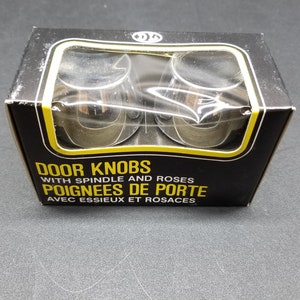May include: A pack of two silver door knobs with a black background and yellow text that reads "DOOR KNOBS WITH SPINDLE AND ROSES POIGNEES DE PORTE AVEC ESSIEUX ET ROSACES".