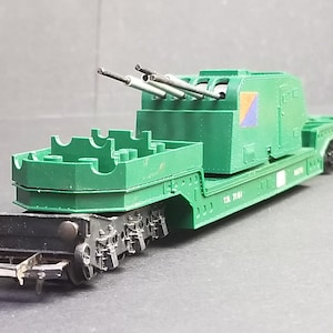 May include: A green model train car with a gun turret and three barrels. The model has a flatbed design with a detailed undercarriage and is painted in a matte green color. The model has a small emblem on the side.