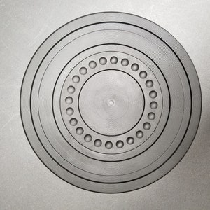 May include: A circular, grey object with concentric rings and a series of evenly spaced holes. The design features a central point surrounded by multiple circular bands, creating a textured surface. The object is set against a grey background.