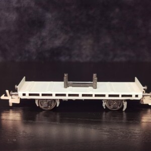 以下が含まれることがあります： 黒い金属製の支柱と車輪が付いた白い模型列車のフラットベッドカー。車は黒い表面にあります。