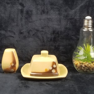 May include: A set of ceramic kitchenware in a yellow and brown color scheme with a speckled pattern. The set includes a butter dish with a lid, a salt shaker, and a small plate. The butter dish has a square shape and the salt shaker is round. The plate is round and has a raised edge. The set is on a black background.