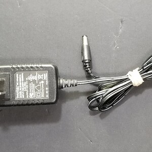 Puede incluir: Adaptador de CA negro con un cable de alimentación negro y un pequeño enchufe negro en el extremo. El adaptador tiene texto que dice "Modelo: Adaptador de CA Entrada: 120V 60Hz 0.3A Salida: 12V 1.5A".