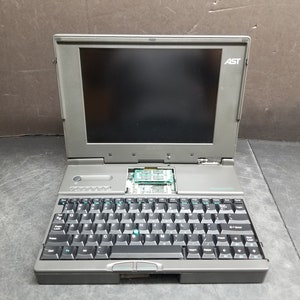 Könnte beinhalten: Ein grauer Laptop mit einer schwarzen Tastatur und einem schwarzen Trackpad. Der Laptop hat einen schwarzen Bildschirm mit dem Wort "AST" in weißen Buchstaben in der oberen rechten Ecke.
