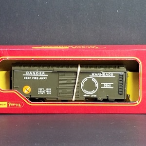 Op de afbeelding: Een groene modeltrein goederenwagon met de woorden "Danger Keep Fire Away" en "Warheads 9841" erop gedrukt. De goederenwagon zit in een rode doos met de woorden "The Gun Box" erop gedrukt.