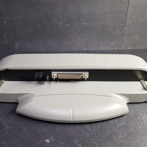 May include: A grey laptop docking station with a black panel featuring a power connector and a data port.