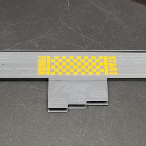 May include: A gray plastic track section for a toy race car set. The section features a checkered flag finish line with the words "START" and "FINISH" printed on either side. The track section has three connection points labeled "CAR B BLACK PLUG", "CAR A BLACK PLUG", and "POWER RED PLUG".