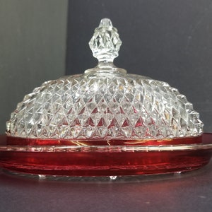 Puede incluir: Un plat de mantequilla de vidrio vintage con una tapa transparente con un patrón de diamante y una base de color rojo intenso.