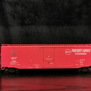 Może przedstawiać: Czerwony model wagonu towarowego z czarnymi kołami. Wagon ma biały napis "Katy MKT 1789" i "Freight-Saver Cushioned". Wagon jest miniaturową repliką prawdziwego wagonu towarowego.