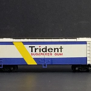May include: A white and blue boxcar with a yellow diagonal stripe and the words "Trident Sugarless Gum" printed on the side.