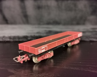 Op de afbeelding: Een rode speelgoedtreinwagen met zwarte wielen en een zwart metalen frame. De wagen heeft een vlakke laadbak met een zwart oppervlak en een wit label met de tekst "SNCF 17423 821504".