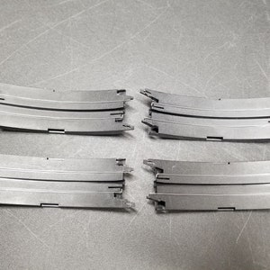 Puede incluir: Cuatro piezas de pista de plástico negro para un circuito de coches de carreras. Las piezas son curvas y tienen tres carriles.