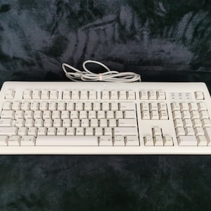 Puede incluir: Un teclado de computadora blanco con una disposición estándar y un teclado numérico. El teclado tiene un cable blanco.