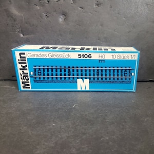 Puede incluir: Una caja azul con texto blanco que dice "Marklin Gerades Gleisstück 5106 HO 10 Stück 1/1". La caja muestra una imagen de una vía de tren.