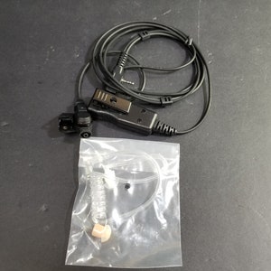 Könnte beinhalten: Ein schwarzes und transparentes Ohrstück mit einem gewickelten Kabel und einem Clip. Das Ohrstück befindet sich in einer durchsichtigen Plastiktüte.