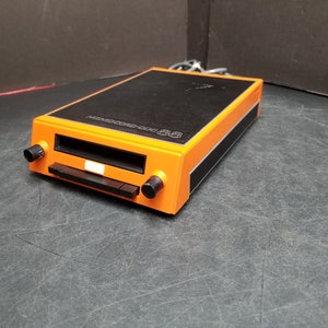 Può includere: Un lettore di cassette arancione e nero con la scritta "MEMOCORD 88" sulla parte superiore.