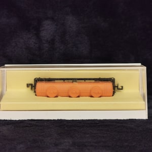 May include: A small, orange model train car in a clear plastic display case. The car has three black wheels and a black metal frame.