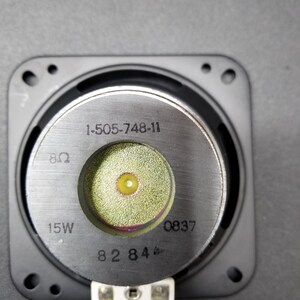 Pu&ograve; includere: Primo piano di un altoparlante nero con un cono di altoparlante argentato. Il cono dell'altoparlante ha il testo "1-505-748-11", "8&Omega;", "15W", "0837" e "82 84" stampato su di esso.