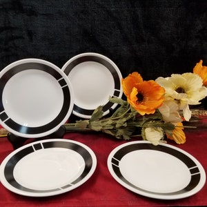 Puede incluir: Cuatro platos de cerámica blanca con ribete negro. Los platos tienen un diseño sencillo con una banda negra alrededor del borde y una raya negra en el centro. Los platos están dispuestos en semicírculo sobre una superficie roja.