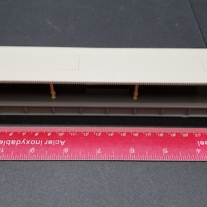 Puede incluir: Una maqueta de andén de estación de tren con techo beige claro y base gris. La plataforma está sostenida por pequeñas columnas amarillas. Una regla roja con pulgadas y centímetros está al lado.
