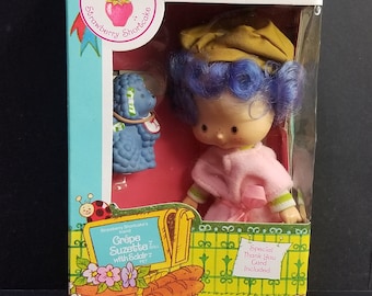 May include: A Strawberry Shortcake doll with a blue poodle pet. The doll is wearing a pink dress with a green trim and a brown hat. The doll and pet are in a box with a yellow and green background. The box says "Strawberry Shortcake's Grêpe Suzette doll with Eclair pet".