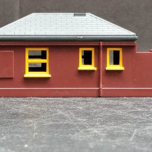 May include: A small, red, plastic model building with a grey roof and three yellow windows. The building has a small, red, plastic wall attached to the side.