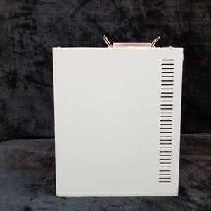 Pode incluir: Uma caixa de metal branca com uma série de pequenos orifícios no lado direito. A caixa tem uma trava de metal na parte superior.