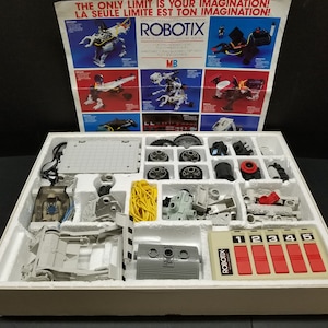 May include: A white box with a grey foam insert containing a robotic construction kit. The box lid is open and shows a colourful instruction sheet with the text "ROBOTIX" and images of different robots. The kit includes a control panel with five red buttons labelled 1 through 5, a grey base, wheels, gears, and other parts.
