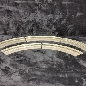 May include: Six curved pieces of track for a model train set. The track is made of a light gray plastic and has a silver metal strip running along the center.