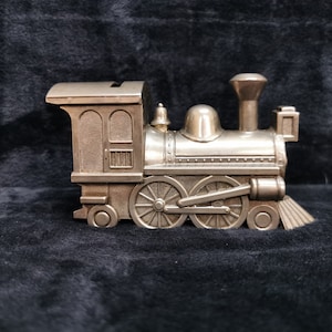 May include: A silver toy train engine against a black background. The train has a bell on top and a slot in the back for coins.