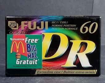 以下が含まれることがあります： 黒いカセットテープに、金色の富士のロゴと黄色の「DR 60」の文字が書かれています。テープには、緑と赤のステッカーが貼られており、「Free Big Mac Gratuit」の文字とマクドナルドのロゴが書かれています。