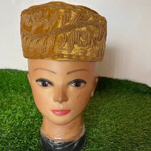May include: A gold-colored, velvet-textured hat with intricate embroidered leaf patterns. The hat is displayed on a mannequin head with a neutral skin tone, set against a green background.