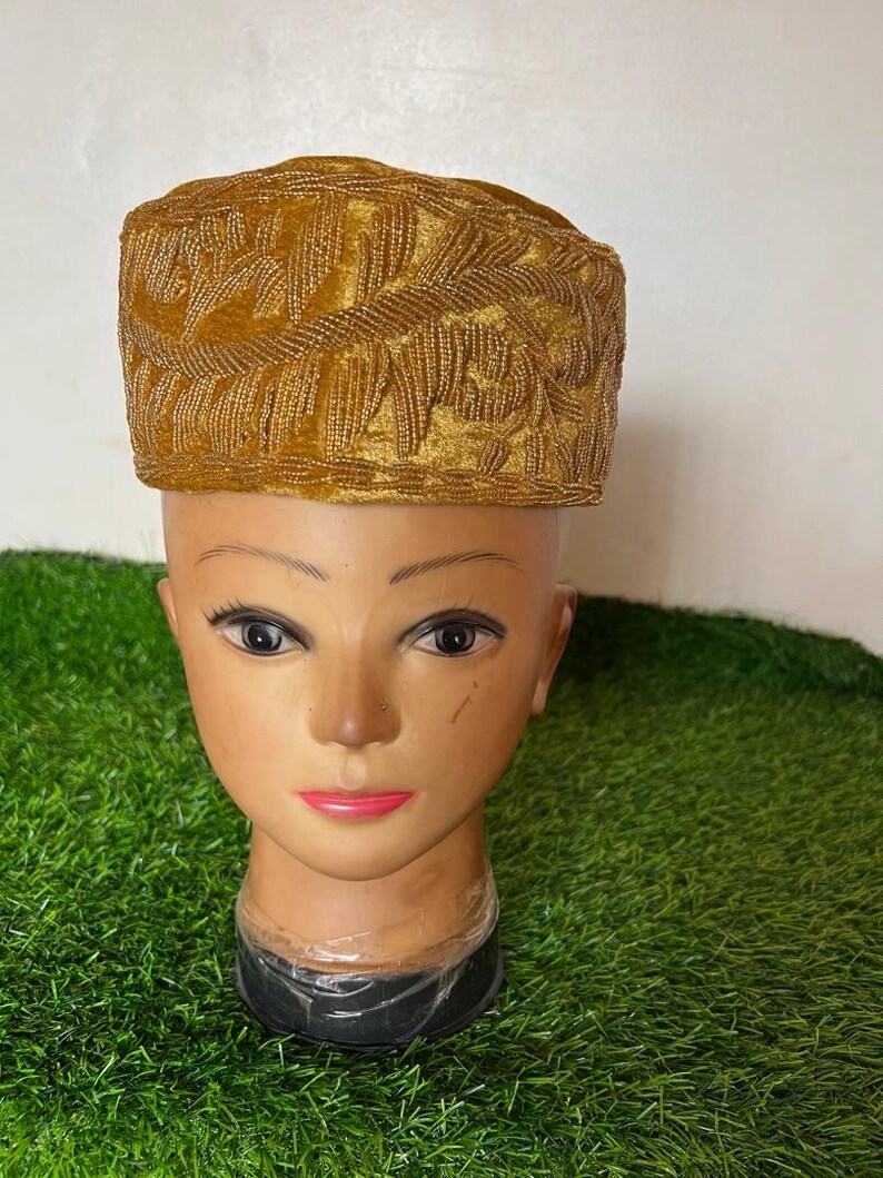 May include: A gold-colored, velvet-textured hat with intricate embroidered leaf patterns. The hat is displayed on a mannequin head with a neutral skin tone, set against a green background.