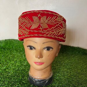 May include: A red velvet hat with gold beaded embroidery. The hat is rectangular with a flat top and a slightly curved brim. The gold embroidery forms a floral pattern. The hat is displayed on a mannequin head.