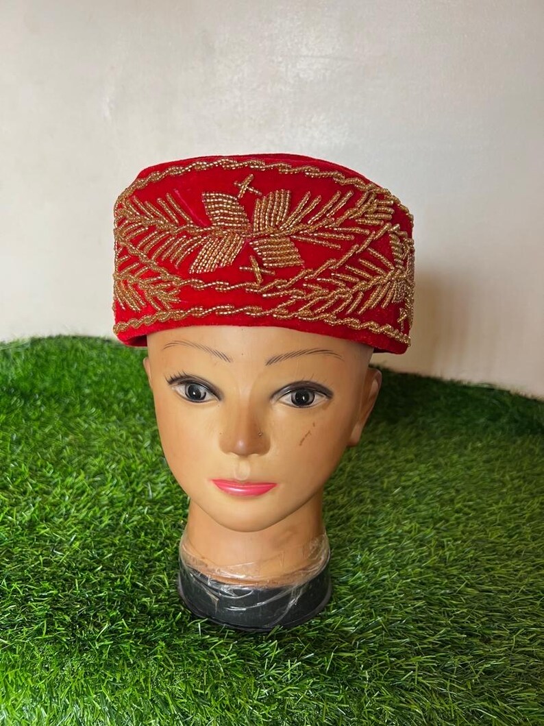 May include: A red velvet hat with gold beaded embroidery. The hat is rectangular with a flat top and a slightly curved brim. The gold embroidery forms a floral pattern. The hat is displayed on a mannequin head.