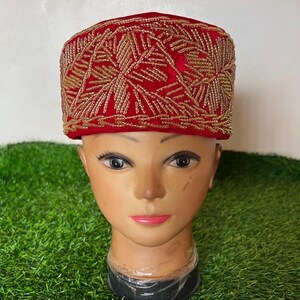 May include: A red velvet cap with intricate gold beaded embroidery. The cap is displayed on a mannequin head against a backdrop of green artificial grass. The gold beading forms a geometric pattern.