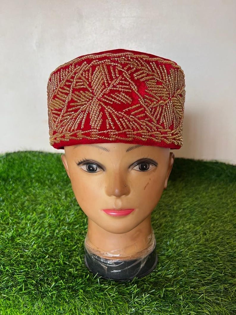 May include: A red velvet cap with intricate gold beaded embroidery. The cap is displayed on a mannequin head against a backdrop of green artificial grass. The gold beading forms a geometric pattern.