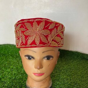 May include: A red hat with gold floral embroidery. The hat is displayed on a mannequin head. The gold embroidery is in the shape of flowers and leaves. The hat is a vibrant red color.