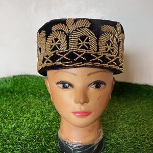 May include: A black velvet hat with intricate gold beaded embroidery. The hat features a rectangular shape with a flat top and a detailed pattern along the brim and crown. The hat is displayed on a mannequin head.