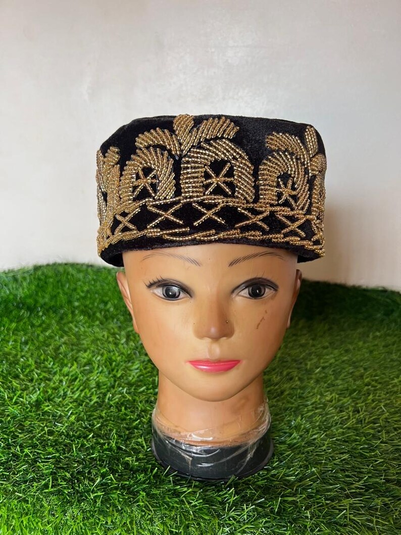 May include: A black velvet hat with intricate gold beaded embroidery. The hat features a rectangular shape with a flat top and a detailed pattern along the brim and crown. The hat is displayed on a mannequin head.