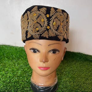 May include: A black velvet hat with intricate gold beaded embroidery. The design features a repeating pattern of stylized leaves and small, round gold accents. The hat is displayed on a mannequin head.
