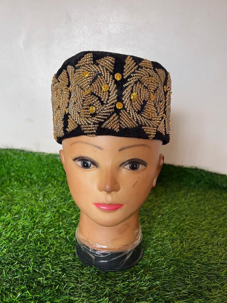 May include: A black velvet hat with intricate gold beaded embroidery. The design features a repeating pattern of stylized leaves and small, round gold accents. The hat is displayed on a mannequin head.