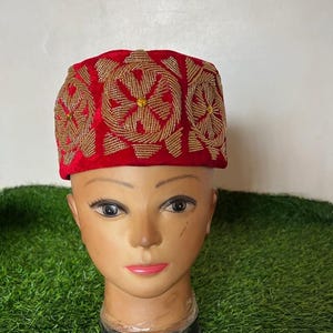 May include: A red velvet hat with intricate gold beaded embroidery. The hat features a rectangular shape with floral patterns. The hat is displayed on a mannequin head. The background is green artificial grass.