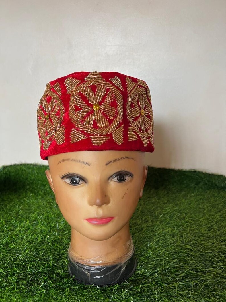May include: A red velvet hat with intricate gold beaded embroidery. The hat features a rectangular shape with floral patterns. The hat is displayed on a mannequin head. The background is green artificial grass.