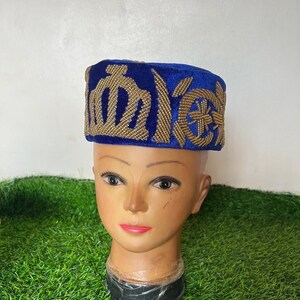 May include: A blue velvet hat with gold embroidered designs. The hat is placed on a mannequin head with a neutral skin tone. The hat's design features geometric and stylized floral patterns. The background is a mix of green and white.